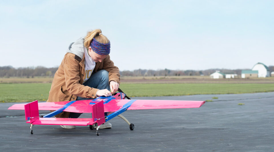 A student kneels near her team's red and blue plane on the airstrip