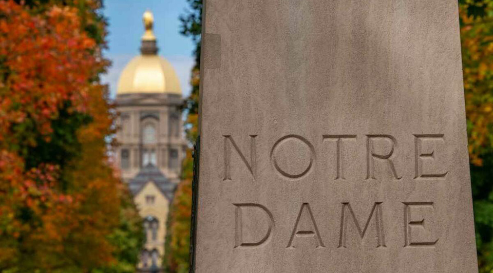 A photo of a stone that reads "Notre Dame" with the main building and autumn trees in the background