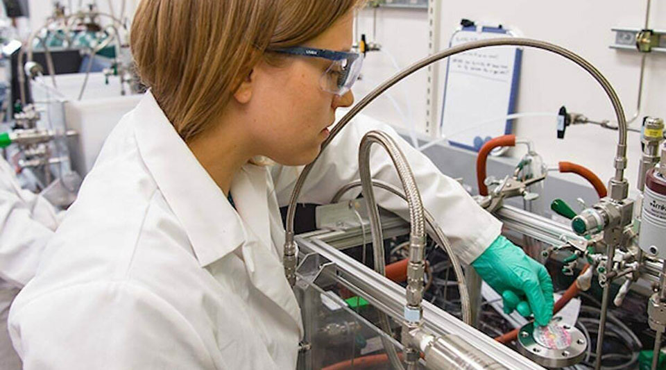 A researcher in a white coat reaches behind a system of wires while holding a round metal device.