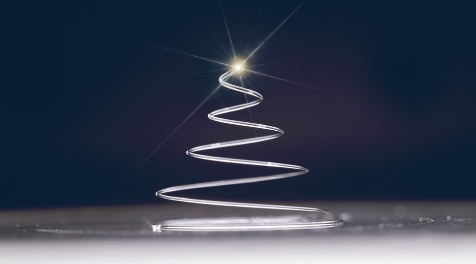 A tightly coiled, spiral-shaped glass Christmas tree created using 3D printing rests on a flat surface. The clear, intricate design is showcased under soft lighting, emphasizing its delicate structure and precision.