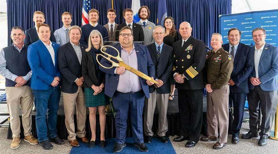 A group of men and women pose for a photo. A person in the center holds large, gold ceremonial scissors, suggesting a ribbon-cutting ceremony. They stand on a blue carpet in front of a navy backdrop with flags and a banner that says "Naval Surface Warfare Center."