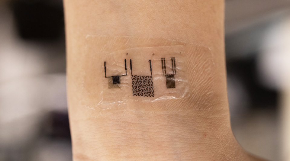 A thin, stretchable bioelectronic device adhered to a wrist, featuring intricate black circuitry patterns on a transparent film.