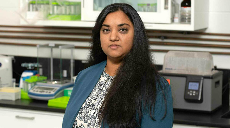 Meenal Datta, a woman with long black hair, wearing a blue blazer and patterned blouse, stands in a laboratory with scientific equipment and glassware in the background. She has a confident and professional expression.